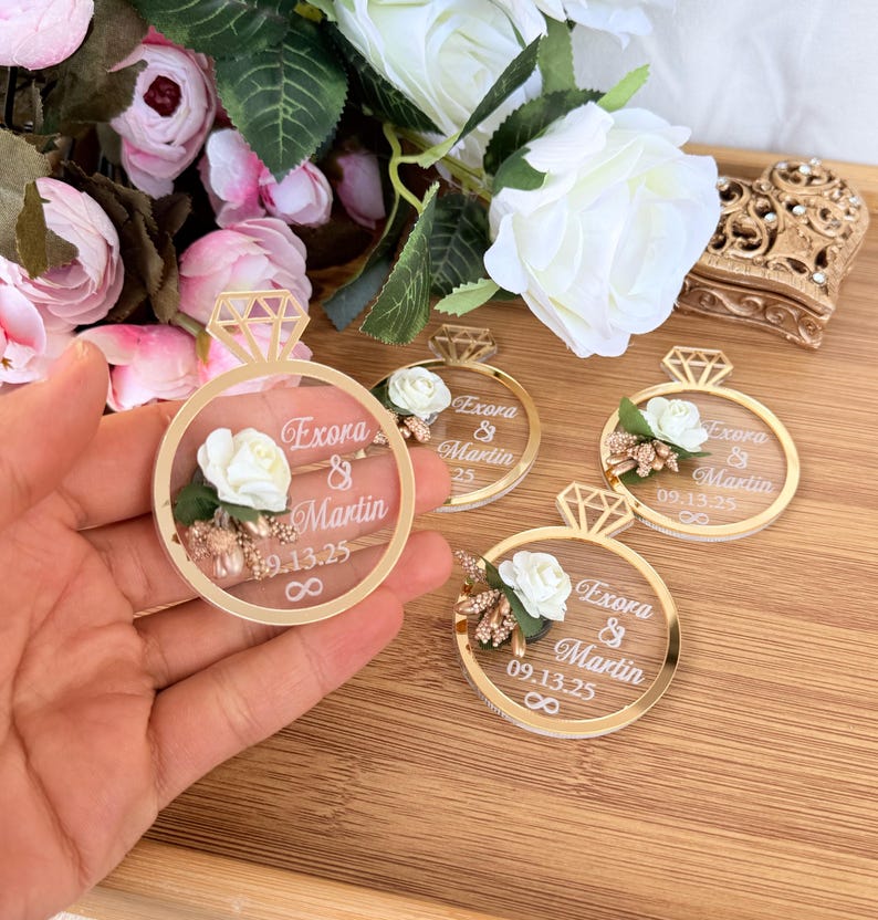 May include: Clear acrylic wedding favors shaped like rings with gold accents. Each ring features a white rose, gold floral details, and the names "Exora & Martin" with the date "09.13.25". The infinity symbol is also included.