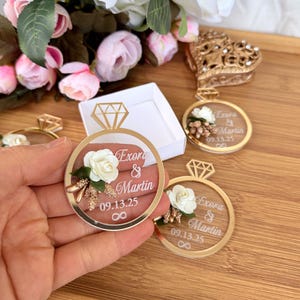 May include: Gold-colored ring-shaped wedding favors with a clear center, featuring a white rose and the names "Exora & Martin" with the date "09.13.25". The favors have a diamond-shaped top and an infinity symbol.