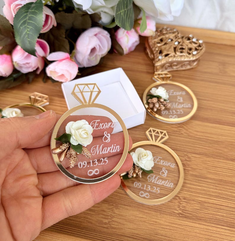 May include: Gold-colored ring-shaped wedding favors with a clear center, featuring a white rose and the names "Exora & Martin" with the date "09.13.25". The favors have a diamond-shaped top and an infinity symbol.