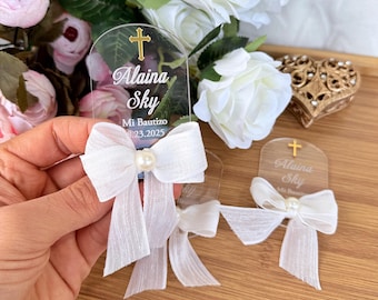 May include: Clear acrylic baptism favors with a gold cross and the name "Alaina Sky" printed on them. Each favor has a white bow with a pearl in the center. The date "23.2025" is also printed on the favors.