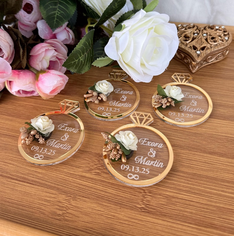 May include: Four gold-rimmed ring-shaped wedding favors with a diamond-shaped top, each featuring a white rose and the names "Exora & Martin" with the date "09.13.25". The favors are on a wooden surface, with flowers in the background.