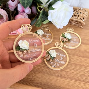May include: Gold-colored ring-shaped wedding favors with a diamond-shaped top. Each favor features a white rose, text that reads "Exora & Martin 09.13.25", and an infinity symbol. The favors are on a wooden surface.