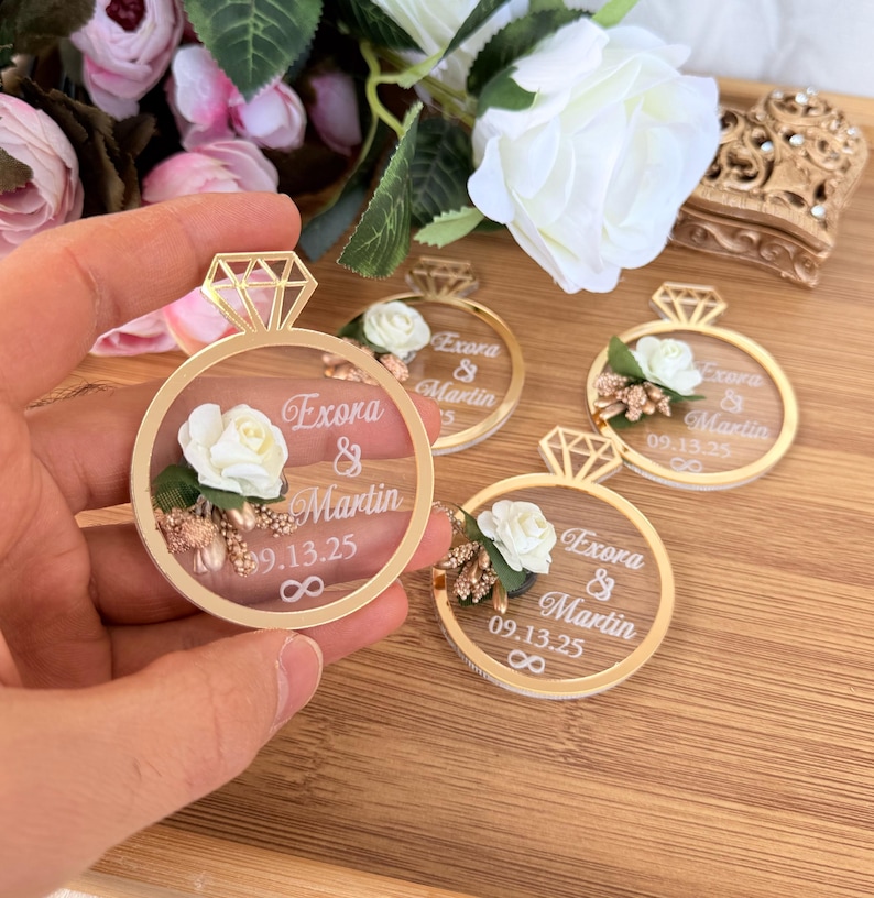 May include: Gold-colored ring-shaped wedding favors with a diamond-shaped top. Each favor features a white rose, text that reads "Exora & Martin 09.13.25", and an infinity symbol. The favors are on a wooden surface.