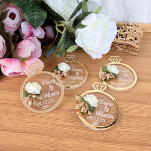 May include: Four gold-rimmed ring-shaped wedding favors with a diamond-shaped top. Each ring features a white rose, gold accents, and the names "Exora & Martin" with the date "09.13.25". The favors are on a wooden surface, with flowers in the background.
