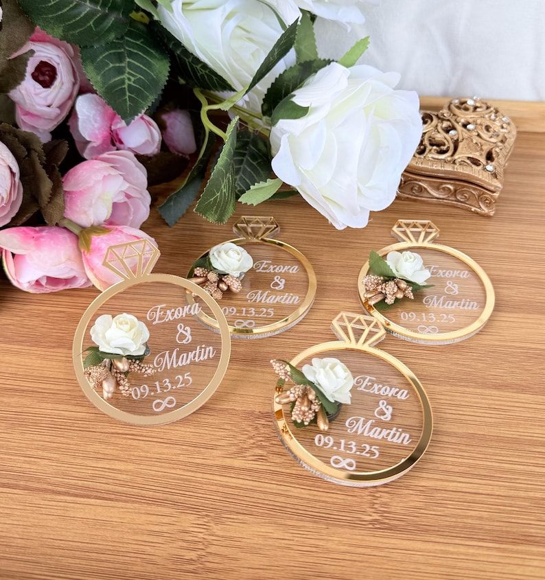 May include: Four gold-rimmed ring-shaped wedding favors with a diamond-shaped top. Each ring features a white rose, gold accents, and the names "Exora & Martin" with the date "09.13.25". The favors are on a wooden surface, with flowers in the background.