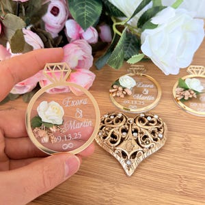 May include: A gold-colored ring-shaped wedding favor with a diamond-shaped top, featuring the names "Exora & Martin" and the date "09.13.25". A gold heart-shaped ornament with intricate details and clear gemstones is also visible.
