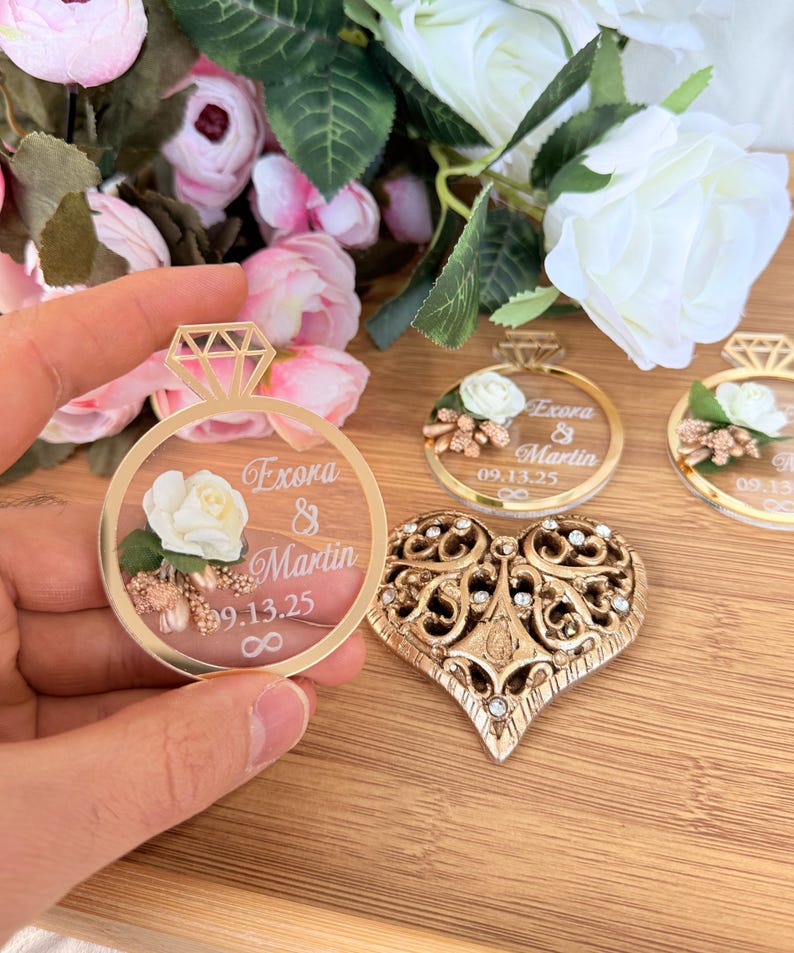 May include: A gold-colored ring-shaped wedding favor with a diamond-shaped top, featuring the names "Exora & Martin" and the date "09.13.25". A gold heart-shaped ornament with intricate details and clear gemstones is also visible.