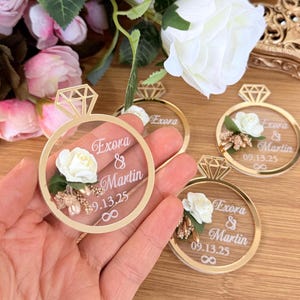 May include: Gold-colored acrylic ring-shaped wedding favors with a diamond-shaped top. Each ring features a white rose, floral accents, and the names "Exora & Martin" with the date "09.13.25".