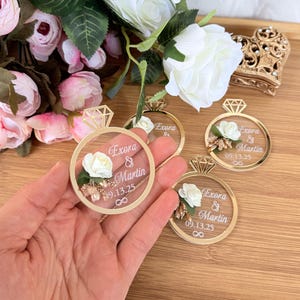 May include: Gold-colored ring-shaped wedding favors with a clear center, featuring a white rose, small gold accents, and the names "Exora & Martin" with a date. The favors are held in a hand, with flowers in the background.