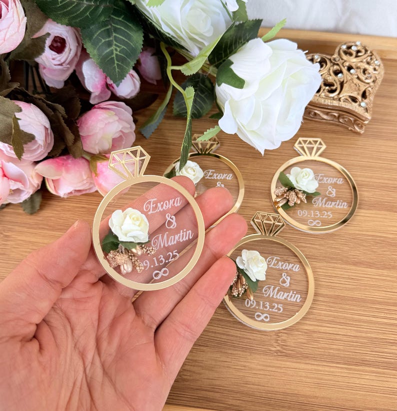 May include: Gold-colored ring-shaped wedding favors with a clear center, featuring a white rose, small gold accents, and the names "Exora & Martin" with a date. The favors are held in a hand, with flowers in the background.