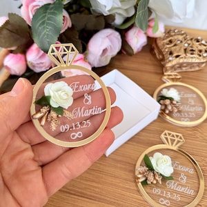 May include: Gold-colored ring-shaped wedding favors with a diamond-shaped top. Each ring features a white rose, text that reads "Exora & Martin 09.13.25" and an infinity symbol. The favors are displayed with a white box and flowers.