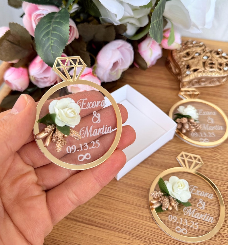 May include: Gold-colored ring-shaped wedding favors with a diamond-shaped top. Each ring features a white rose, text that reads "Exora & Martin 09.13.25" and an infinity symbol. The favors are displayed with a white box and flowers.