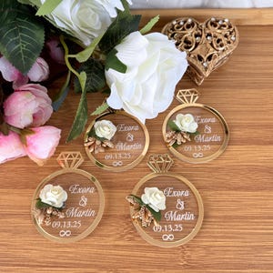 May include: Gold-colored acrylic ring-shaped wedding favors with a diamond-shaped top, featuring the names "Exora & Martin" and the date "09.13.25". Each ring is adorned with a white rose and floral accents. The favors are arranged on a wooden surface.