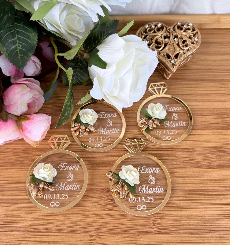 May include: Gold-colored acrylic ring-shaped wedding favors with a diamond-shaped top, featuring the names "Exora & Martin" and the date "09.13.25". Each ring is adorned with a white rose and floral accents. The favors are arranged on a wooden surface.