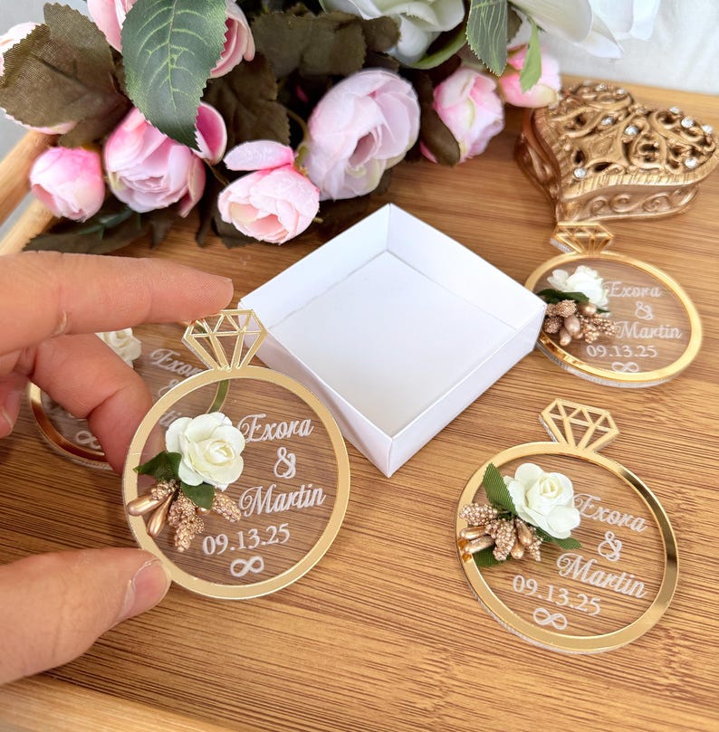 May include: Clear acrylic ring-shaped wedding favors with gold accents, featuring a diamond-shaped top and floral embellishments. Each ring displays the names "Exora & Martin", date "09.13.25", and an infinity symbol. A white box and pink flowers are in the background.