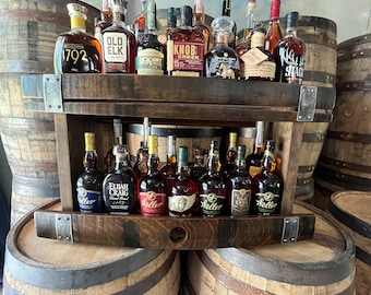 May include: A wooden shelf unit with two tiers, made from reclaimed whiskey barrels, holds a collection of various bourbon and whiskey bottles. The bottles are arranged on the shelves, with some bottles on the top shelf and others on the bottom shelf. The shelf unit is made of dark wood and has a rustic appearance.