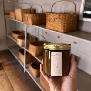 May include: A brown glass jar candle with a gold lid and a white label that reads Stinson Provisions Basil and Bergamot. The candle is held in front of a white shelving unit filled with various woven wicker baskets of different sizes and shapes.