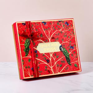 May include: A square red gift box featuring a gold tree illustration, two green peacocks, and a red ribbon tied in a bow. A central gold label displays the text "छप्पन भोग प्रसादम्" in a script font. The box has gold edges and a decorative pattern.