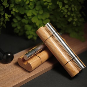 Puede incluir: Un instrumento musical de madera y metal, posiblemente una maza o un instrumento de percusión, apoyado sobre una superficie de madera. El instrumento tiene un cuerpo cilíndrico de madera con detalles metálicos y está asegurado con bandas negras. También se ve una esfera negra.