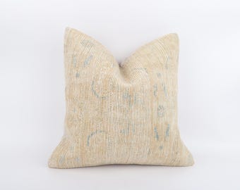 May include: A square cushion in beige with a textured surface and subtle blue and beige patterns. The cushion has a soft appearance, suitable for home decor. The design features a mix of vertical and circular elements.
