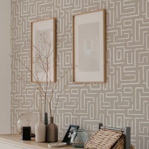 Puede incluir: Dos cuadros enmarcados con un fondo marrón claro cuelgan de una pared con un papel tapiz de patrón geométrico beige y blanco. Un jarrón con ramas secas se encuentra sobre una mesa con un tarro de vidrio, una vela y una cesta de mimbre.