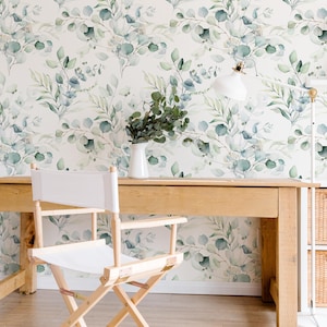May include: A light wood desk with a white director's chair and a white vase with greenery. The background is a wallpaper with a watercolor eucalyptus leaf pattern. A white desk lamp is on the desk.