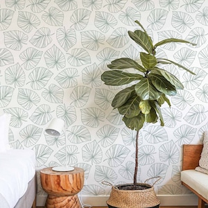 May include: A white wall with a light green leaf pattern wallpaper. A large green plant in a woven basket sits in front of the wall. A wooden side table with a white lamp sits next to the plant. A white bed with a white blanket is in the background.