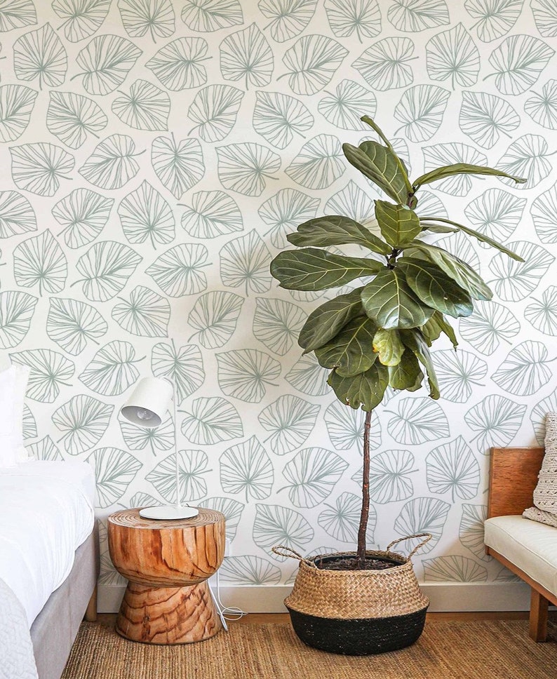 May include: A white wall with a light green leaf pattern wallpaper. A large green plant in a woven basket sits in front of the wall. A wooden side table with a white lamp sits next to the plant. A white bed with a white blanket is in the background.