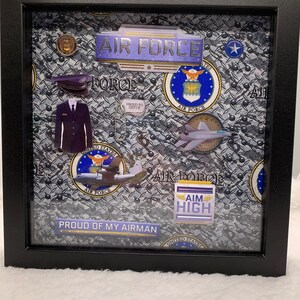 May include: A framed shadow box with a gray and black camouflage pattern. The shadow box includes a variety of Air Force patches, a miniature Air Force uniform, a small airplane, and the text "Proud of My Airman".