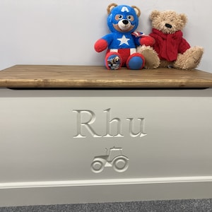 May include: White wooden toy box with a brown lid. The box is engraved with the name "Rhu" and a tractor design. Two stuffed animals, a red and white teddy bear and a blue and red Captain America teddy bear, are sitting on the lid.