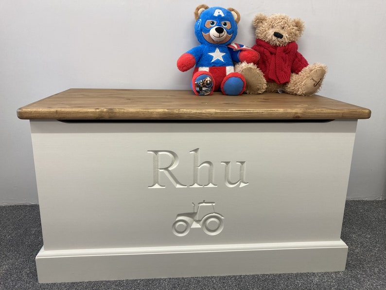 May include: White wooden toy box with a brown lid. The box is engraved with the name "Rhu" and a tractor design. Two stuffed animals, a red and white teddy bear and a blue and red Captain America teddy bear, are sitting on the lid.
