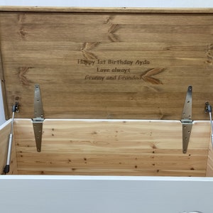 May include: White wooden toy chest with a hinged lid and a brown wooden top. The lid is open and the inside of the chest is visible. The top of the chest is engraved with the text "Happy 1st Birthday Ayda Love always Granny and Grandad".