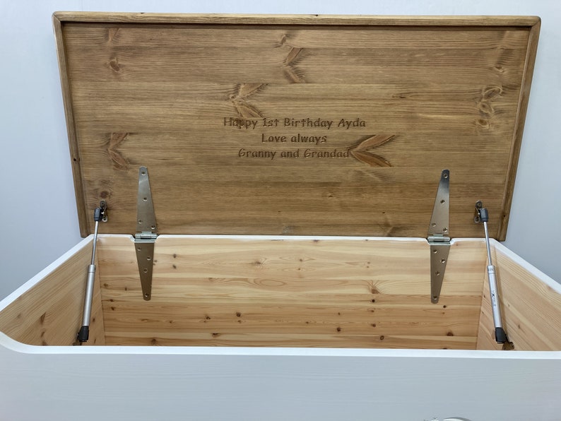 May include: White wooden toy chest with a hinged lid and a brown wooden top. The lid is open and the inside of the chest is visible. The top of the chest is engraved with the text "Happy 1st Birthday Ayda Love always Granny and Grandad".