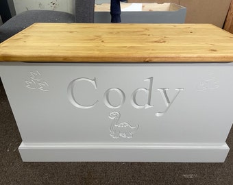 May include: A white wooden toy chest with a light wood top. The chest has a dinosaur and the name "Cody" carved into the front.