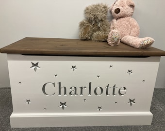 May include: White wooden toy chest with a brown lid. The chest has a silver star design and the name "Charlotte" carved into the front. Two plush teddy bears are sitting on top of the chest.