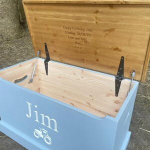 May include: A light blue wooden toy box with a hinged lid. The lid is open and has a personalized message that reads "Happy birthday Jim! 3 today, 31/01/23 Love you lots Mum xxx". The front of the box has the name "Jim" and a tractor graphic.