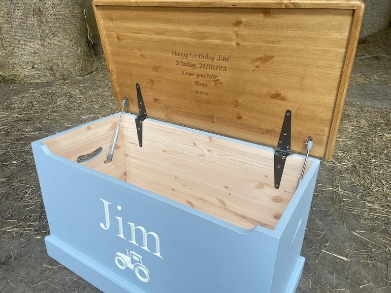 May include: A light blue wooden toy box with a hinged lid. The lid is open and has a personalized message that reads "Happy birthday Jim! 3 today, 31/01/23 Love you lots Mum xxx". The front of the box has the name "Jim" and a tractor graphic.