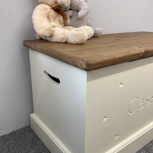 May include: White wooden toy chest with a brown lid. The chest has a cutout handle and is decorated with stars and the name "Cassie". Two stuffed animals, a bear and an elephant, are sitting on the lid.