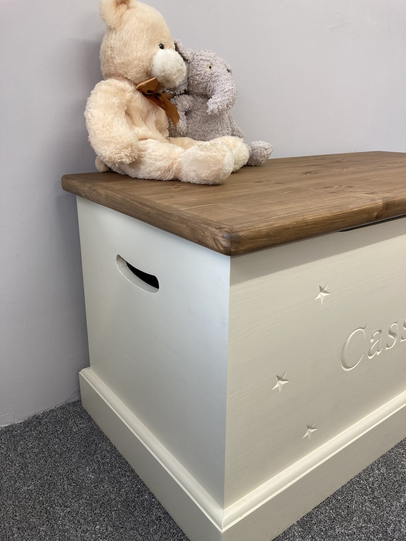 May include: White wooden toy chest with a brown lid. The chest has a cutout handle and is decorated with stars and the name "Cassie". Two stuffed animals, a bear and an elephant, are sitting on the lid.
