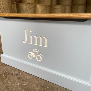 May include: A light blue wooden toy box with a light brown lid. The box has a white tractor and the name "Jim" carved into the front.