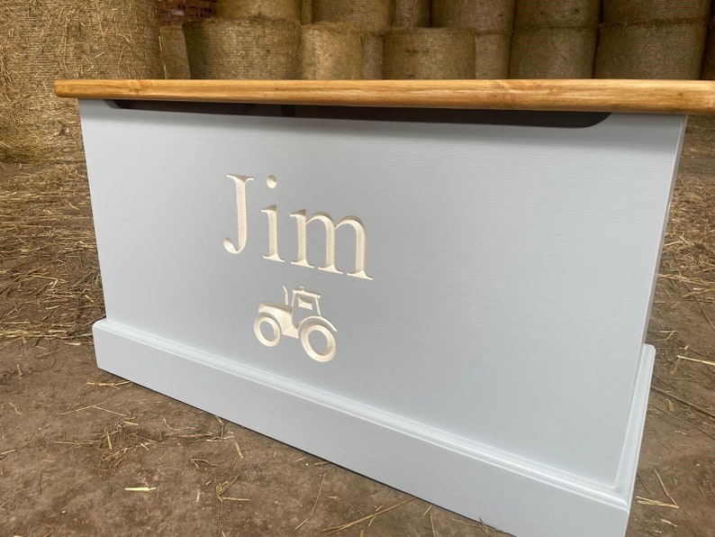May include: A light blue wooden toy box with a light brown lid. The box has a white tractor and the name "Jim" carved into the front.