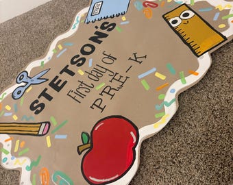 Puede incluir: Un letrero hecho a mano para el primer día de Pre-K, con el nombre "Stetson's". El letrero es marrón con borde blanco e ilustraciones coloridas de útiles escolares, incluyendo una manzana roja, una regla y un lápiz.