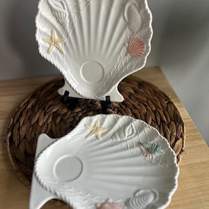 May include: Two white ceramic seashell-shaped serving dishes. Each dish features embossed seashells, starfish, and a net design. The dishes have a central circular indentation and are supported by black stands. The dishes are on a woven mat.