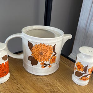Puede incluir: Un juego de tres artículos de cocina de cerámica con un diseño floral. Los artículos son una jarra y dos saleros y pimenteros. El diseño floral presenta flores naranjas y marrones sobre un fondo blanco.