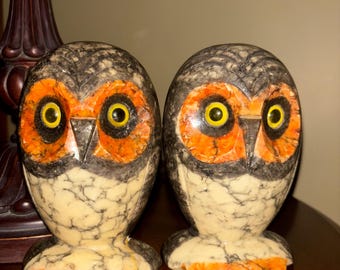 May include: Two owl figurines with a speckled cream and black pattern. The owls have orange and yellow accents on their faces and feet. The eyes are yellow with black pupils. The figurines are displayed on a wooden surface.