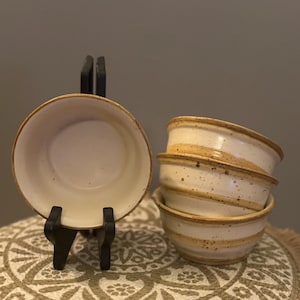 May include: Four off-white ceramic bowls with a speckled finish and a brown rim. One bowl is displayed on a black stand, while the other three are stacked. The bowls are on a patterned, tan surface.