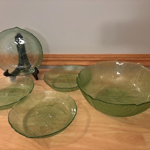 Könnte beinhalten: Ein Set aus grünen Glasschalen in Blattform und einem Teller. Der Teller steht auf einem schwarzen Ständer. Die Schalen und der Teller haben ein strukturiertes Blattdesign. Die Schalen sind in verschiedenen Größen erhältlich.