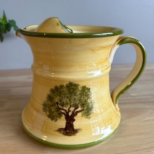 May include: A yellow ceramic pitcher with a green rim and handle. The pitcher features a painted olive tree design.
