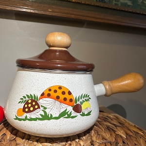 May include: A white speckled enamelware pot with a brown lid and wooden handle. The pot features a colourful mushroom design with orange, yellow, and brown caps. The pot is sitting on a woven surface.