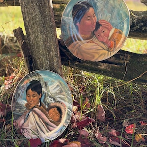 May include: Two round decorative porcelain plates featuring detailed illustrations of individuals in traditional fur-trimmed parkas. One plate shows a person holding a child, while the other depicts a person carrying a child on their back.